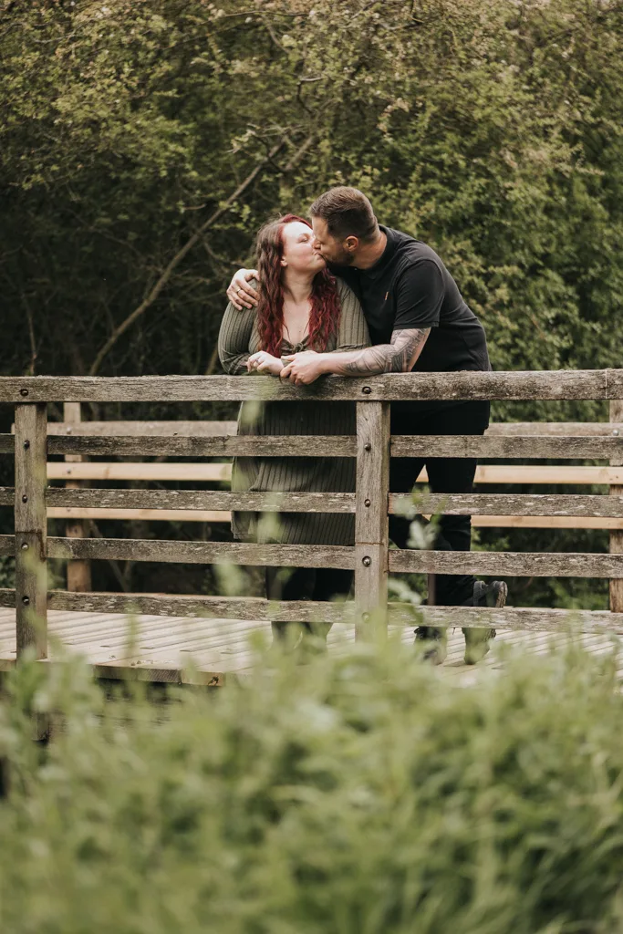 A couple stands on a rustic wooden bridge surrounded by greenery. In this natural outdoor setting in Cleethorpes, the man kisses his partner as they lean on the railing, sharing an affectionate engagement moment. © Aimee Lince Photography - Wedding photographer in Lincolnshire, Yorkshire & Nottinghamshire