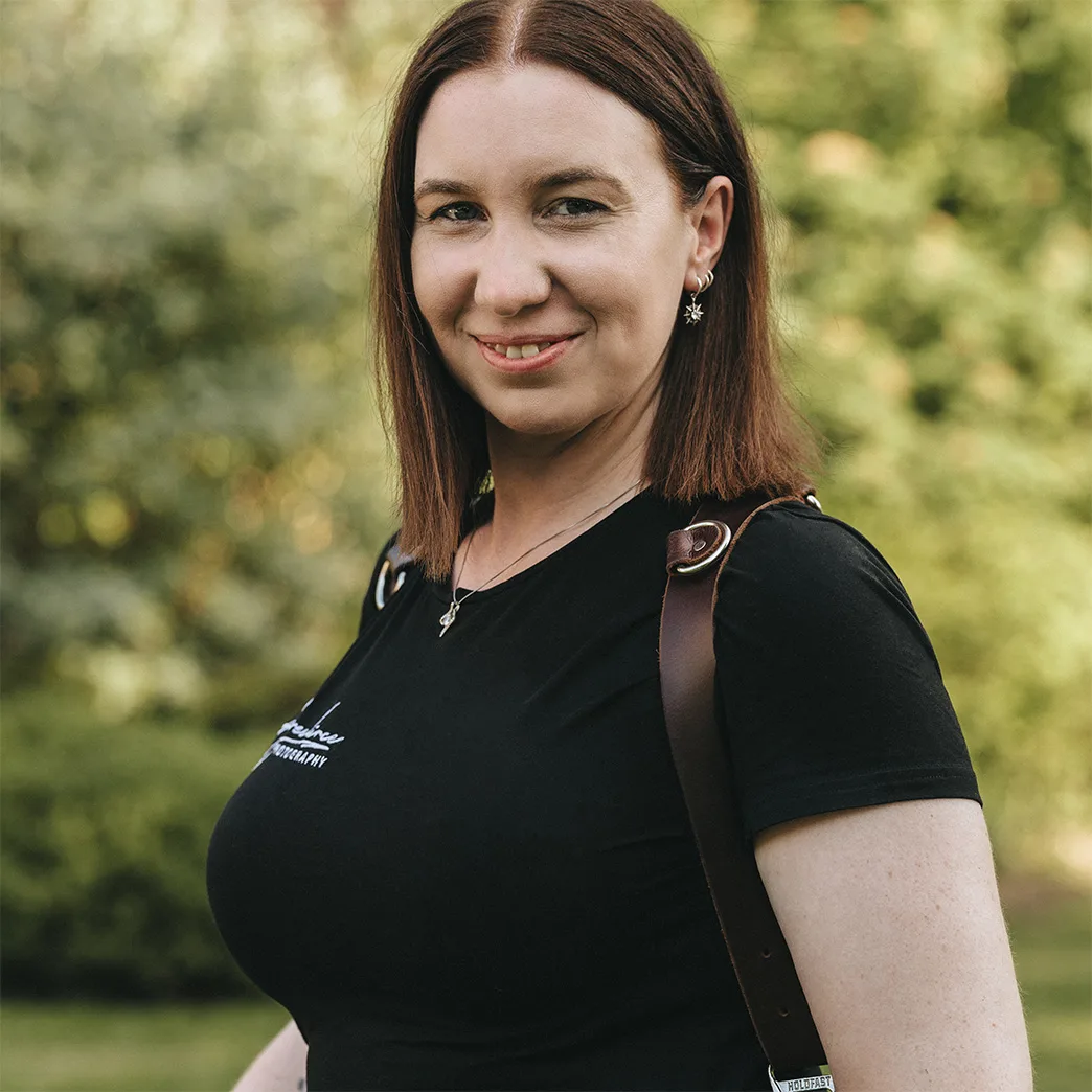 A woman with straight brown hair, wearing a black t-shirt and leather strap over her shoulder, smiles outdoors. She stands in front of blurred green foliage, her natural look complemented by a necklace and small earrings. © Aimee Lince Photography - Wedding photographer in Lincolnshire, Yorkshire & Nottinghamshire