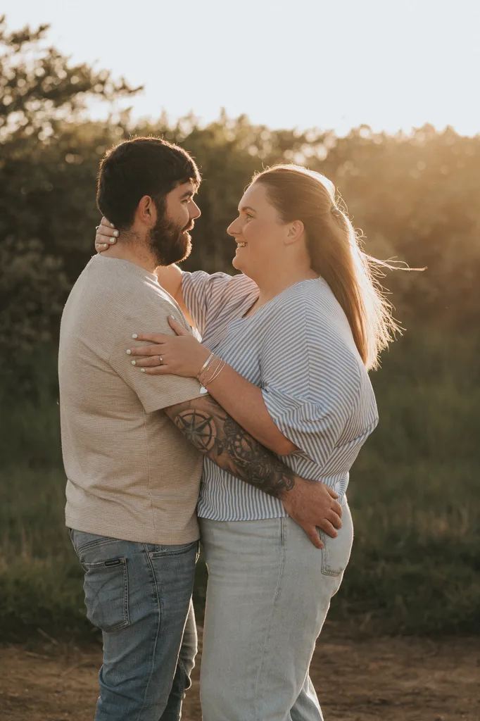 A couple stands outdoors at sunset, smiling at each other in a tender pre wedding moment. The woman wears a striped shirt, while the bearded man with tattooed arms holds her close—capturing their love through engagement photography. © Aimee Lince Photography - Wedding photographer in Lincolnshire, Yorkshire & Nottinghamshire