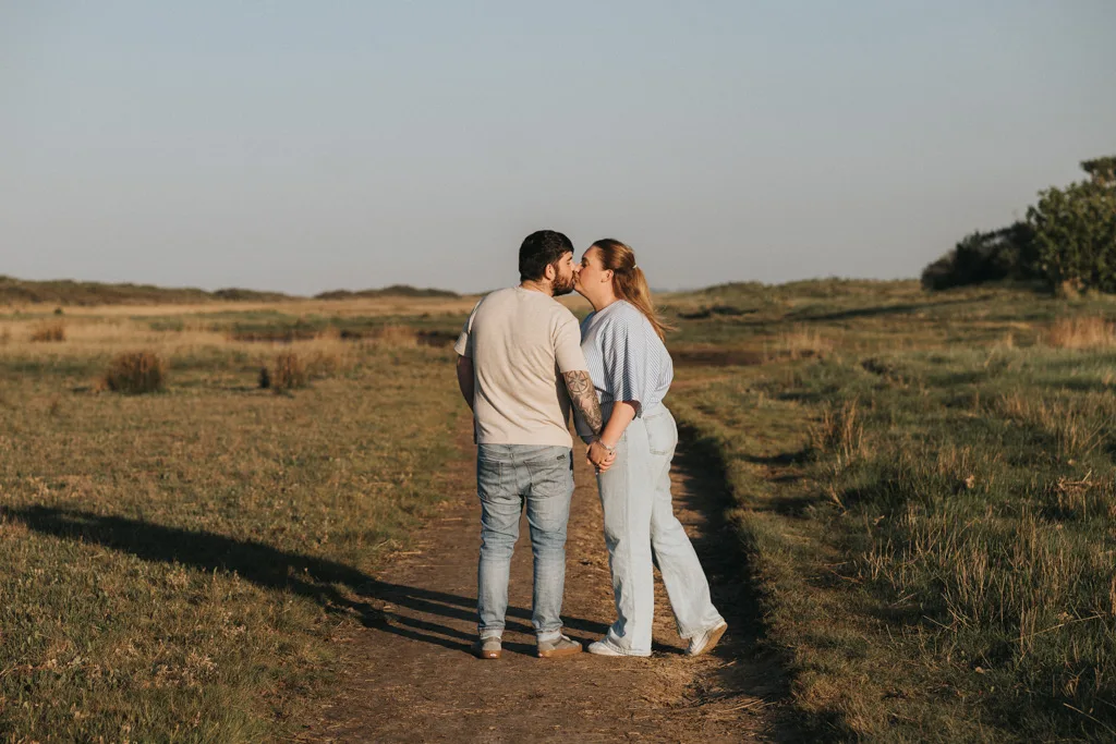 A couple stands on a dirt path in a grassy field near Cleethorpes, holding hands and kissing. The person on the left has short dark hair, tattoos, and casual attire; the right wears a blue shirt—capturing a sweet pre wedding moment. © Aimee Lince Photography - Wedding photographer in Lincolnshire, Yorkshire & Nottinghamshire