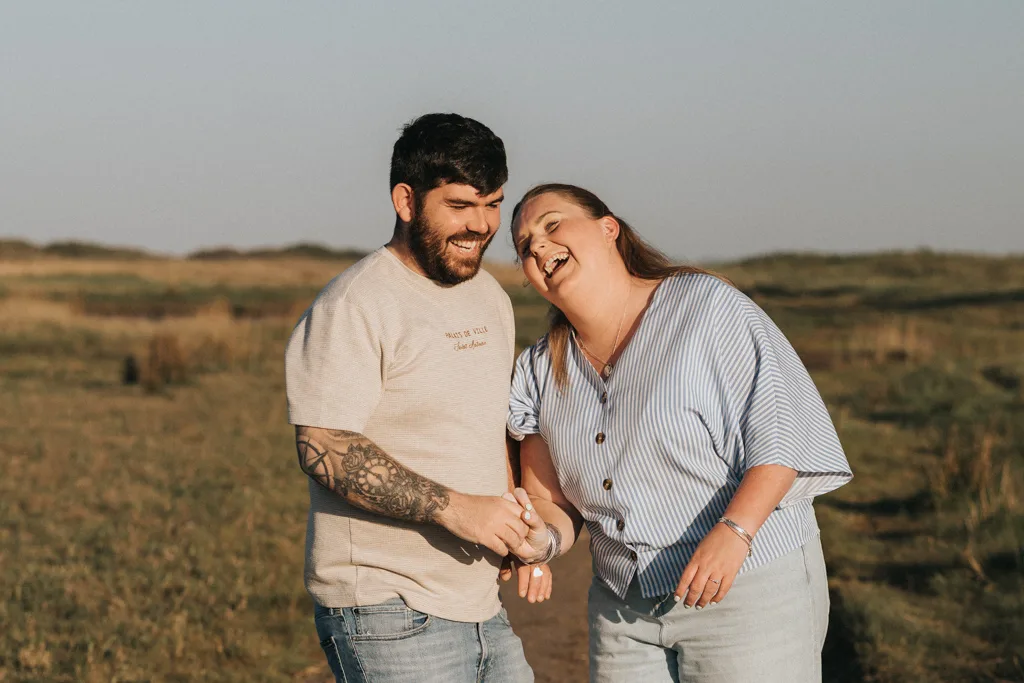 A man and woman laugh joyfully together in a sunny, grassy field—perfect for pre wedding photography. The bearded, tattooed man wears a beige shirt and jeans, while she leans in smiling, dressed in a blue-striped blouse and jeans. © Aimee Lince Photography - Wedding photographer in Lincolnshire, Yorkshire & Nottinghamshire