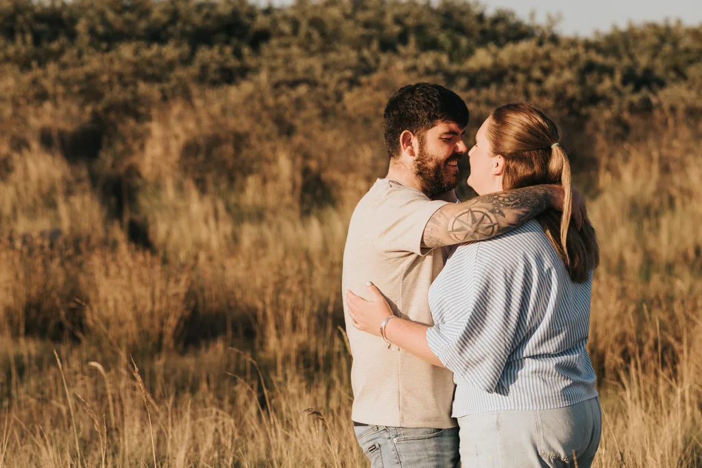 A couple embraces in a field of tall grass, smiling at each other during their engagement photography session. The tattooed man wears a beige shirt, while the woman in a blue and white striped top stands among Cleethorpes' lush bushes and greenery. © Aimee Lince Photography - Wedding photographer in Lincolnshire, Yorkshire & Nottinghamshire