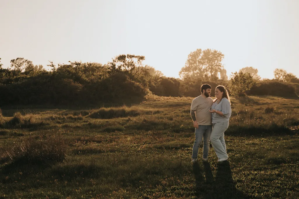 A couple stands close together in a sunlit field near Cleethorpes, smiling at each other. Their casual outfits and the golden glow of sunset make this engagement photography moment feel warm and magical among the green grass and trees. © Aimee Lince Photography - Wedding photographer in Lincolnshire, Yorkshire & Nottinghamshire