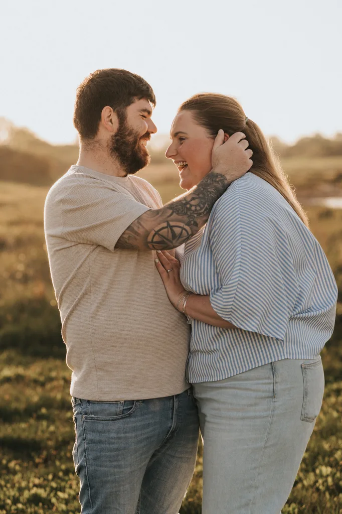 A bearded man with a tattoo on his arm gently touches his fiancée’s face in a sunlit field near Cleethorpes. Both wear light, casual clothes and look happy, sharing an intimate pre wedding moment outdoors. © Aimee Lince Photography - Wedding photographer in Lincolnshire, Yorkshire & Nottinghamshire