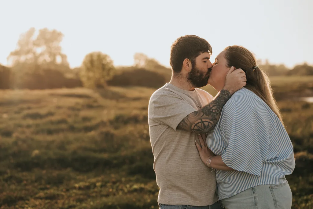 A couple shares a kiss in a grassy Cleethorpes field at sunset. The pre wedding photography captures his tattoos and beard, her striped blouse, and the gentle way she holds his arm, all softly lit by the fading light. © Aimee Lince Photography - Wedding photographer in Lincolnshire, Yorkshire & Nottinghamshire