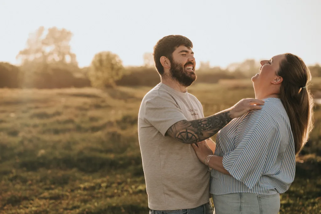Amy & Ethan Engagement Photo Shoot at Cleethorpes Beach and Boating Lake 67 of 81 © Aimee Lince Photography - 12.05.2025 A man and woman stand outdoors in a sunlit field, smiling and laughing together during their pre wedding photography session. The man has a beard and tattoos; the woman wears a striped shirt. Joyful moments unfold with trees and grass in the background. © Aimee Lince Photography - Wedding photographer in Lincolnshire, Yorkshire & Nottinghamshire
