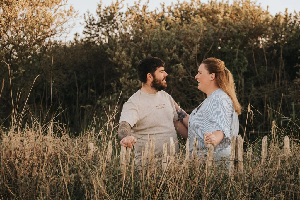 A couple stands outdoors in Cleethorpes, smiling as they hold hands over a rustic wooden fence surrounded by tall grass. Perfect for a pre wedding or engagement shoot, the scene is filled with natural beauty and warm connection. © Aimee Lince Photography - Wedding photographer in Lincolnshire, Yorkshire & Nottinghamshire