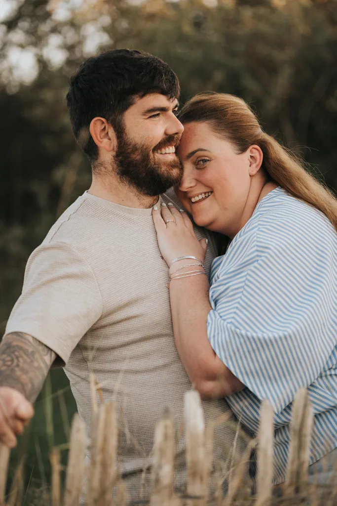 A smiling man with a beard and tattoos hugs his fiancée in a blue and white striped top, her engagement ring on display. This pre wedding photography moment captures their joy outdoors by a rustic wooden fence, with blurred greenery in the background. © Aimee Lince Photography - Wedding photographer in Lincolnshire, Yorkshire & Nottinghamshire