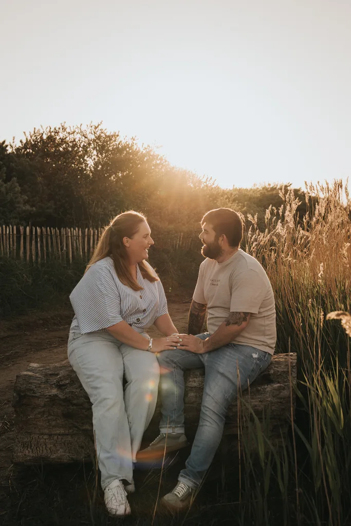 A couple sits on a log outdoors at sunset, facing each other and smiling. Tall grasses and a wooden fence are in the background. Warm sunlight creates a soft, golden glow around this joyful pre wedding photography moment. © Aimee Lince Photography - Wedding photographer in Lincolnshire, Yorkshire & Nottinghamshire