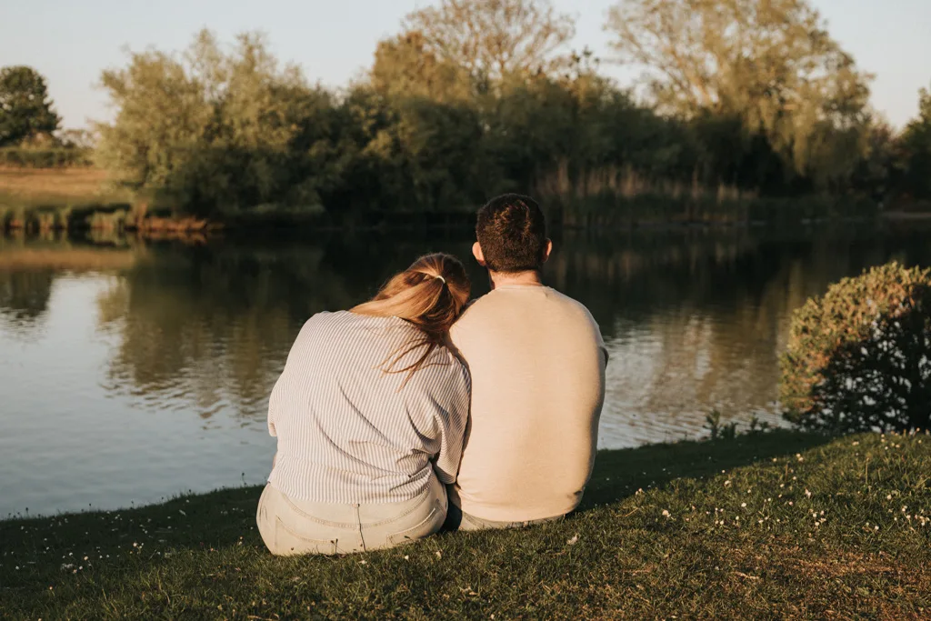 Two people sit on the grass by a calm lake, backs to the camera, sharing a quiet moment. This serene pre wedding photography scene is framed by lush trees and bushes under a clear sky, capturing the essence of a peaceful, sunny day. © Aimee Lince Photography - Wedding photographer in Lincolnshire, Yorkshire & Nottinghamshire