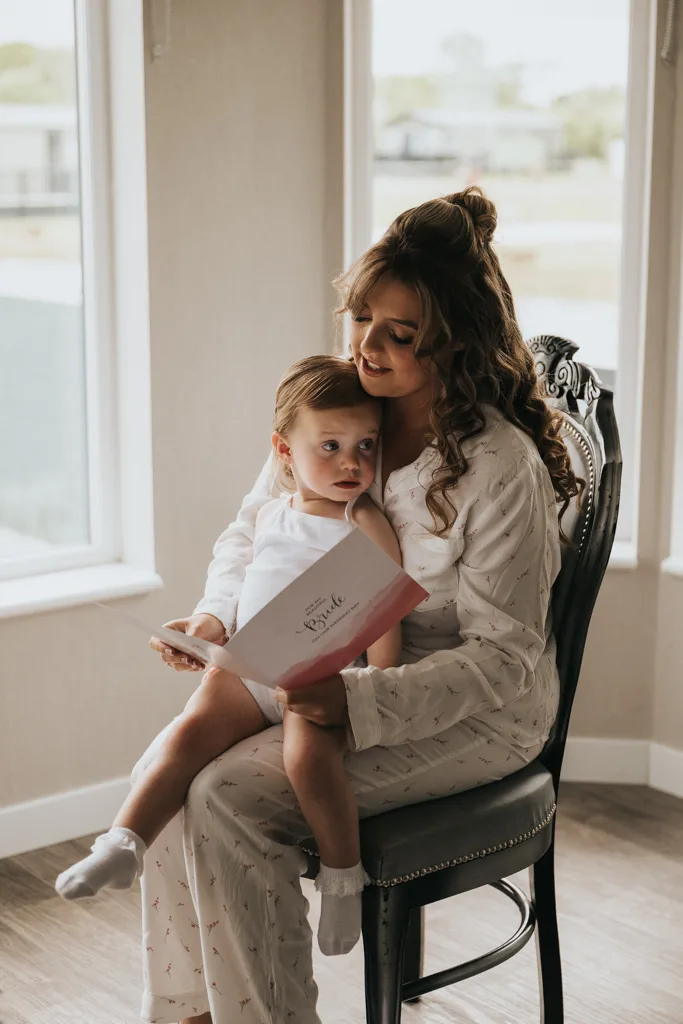 A woman in light pajamas sits on a chair by sunny windows, holding a young child on her lap. Captured in beautiful photography, she reads a book titled "Bible" to the child, creating a warm and nurturing moment. © Aimee Lince Photography - Wedding photographer in Lincolnshire, Yorkshire & Nottinghamshire