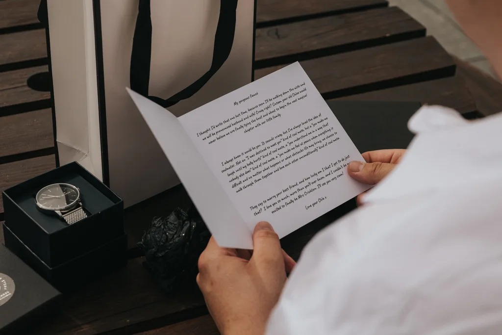 A person in a white shirt sits at a wooden table, reading a letter—likely wedding correspondence. On the table are a wristwatch in an open black box, a decorative item, and a Brackenborough photography gift bag. The person’s face is not visible. © Aimee Lince Photography - Wedding photographer in Lincolnshire, Yorkshire & Nottinghamshire