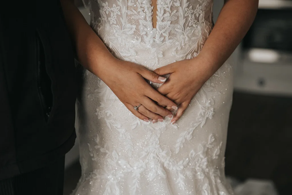 A close-up of a bride’s hands resting gently at her waist, showing a diamond engagement ring. She is wearing a white lace wedding dress with floral patterns—captured beautifully by brackenborough wedding photography—with another person partly visible beside her. © Aimee Lince Photography - Wedding photographer in Lincolnshire, Yorkshire & Nottinghamshire