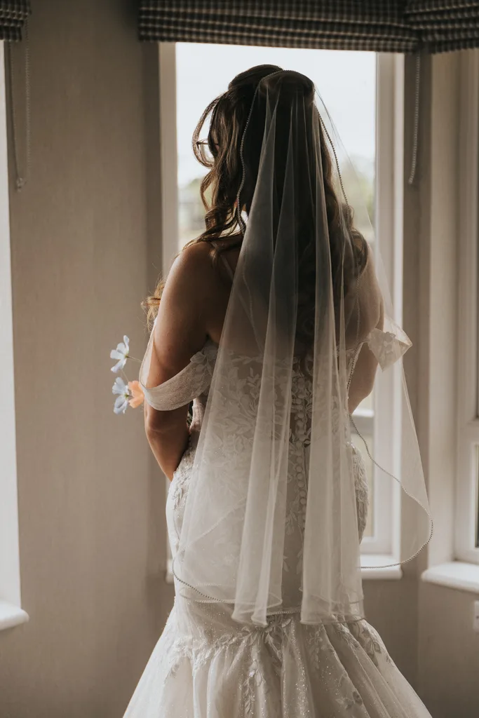 A bride in a white lace off-shoulder gown and sheer veil stands with her back to the camera, gazing out of a sunlit window at Brackenborough. Perfect wedding photography captures her wavy hair and bouquet of white flowers in hand. © Aimee Lince Photography - Wedding photographer in Lincolnshire, Yorkshire & Nottinghamshire