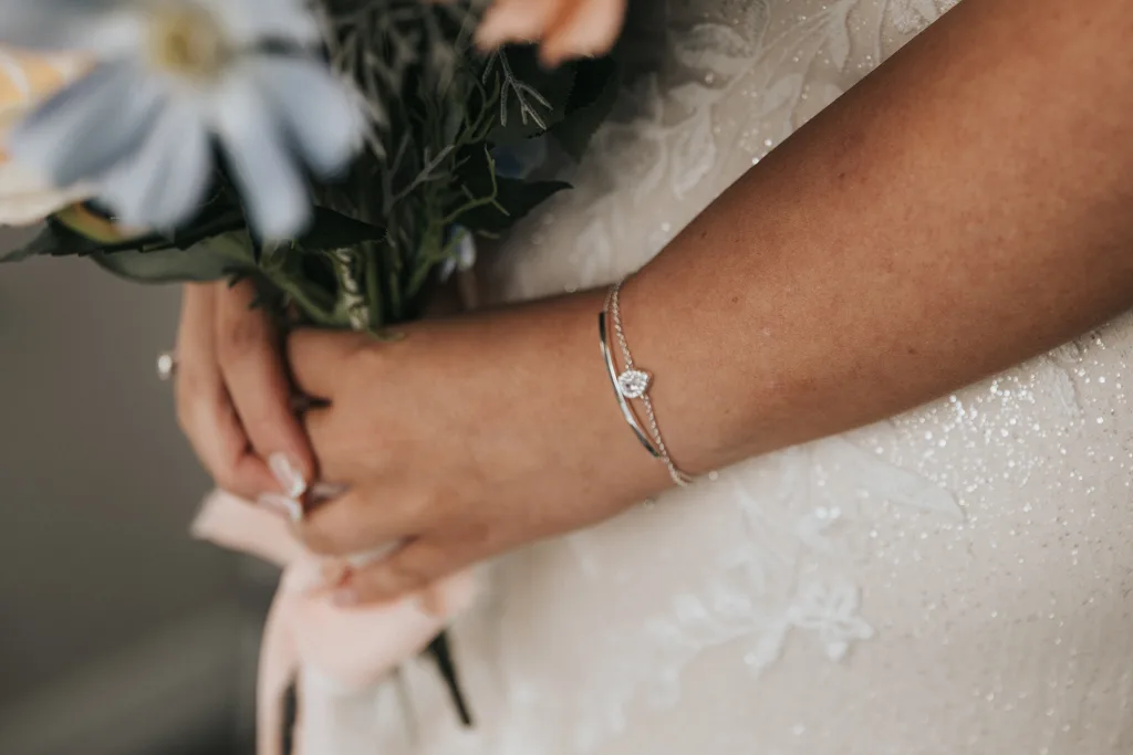 A close-up of a bride’s hands holding a bouquet of flowers. She wears a sparkling bracelet and a simple ring on one finger. Her white dress, with delicate lace details, shines in this elegant moment of wedding photography at Brackenborough. © Aimee Lince Photography - Wedding photographer in Lincolnshire, Yorkshire & Nottinghamshire