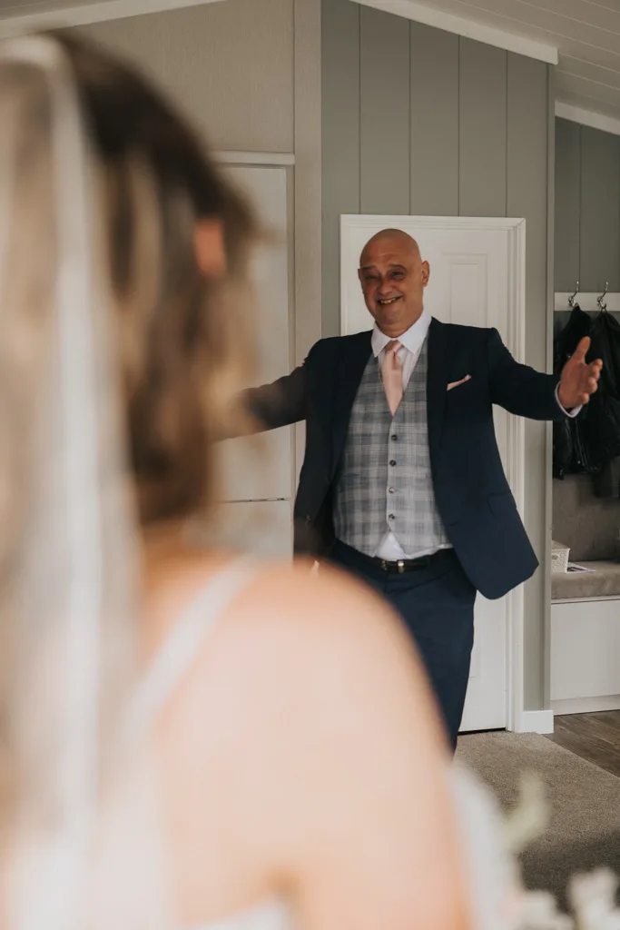 A bald man in a navy suit, checked vest, and pink tie smiles warmly with open arms toward a bride in a gown. Captured indoors at Brackenborough, this wedding photography scene features soft lighting and charming details like a white door and coat hooks. © Aimee Lince Photography - Wedding photographer in Lincolnshire, Yorkshire & Nottinghamshire