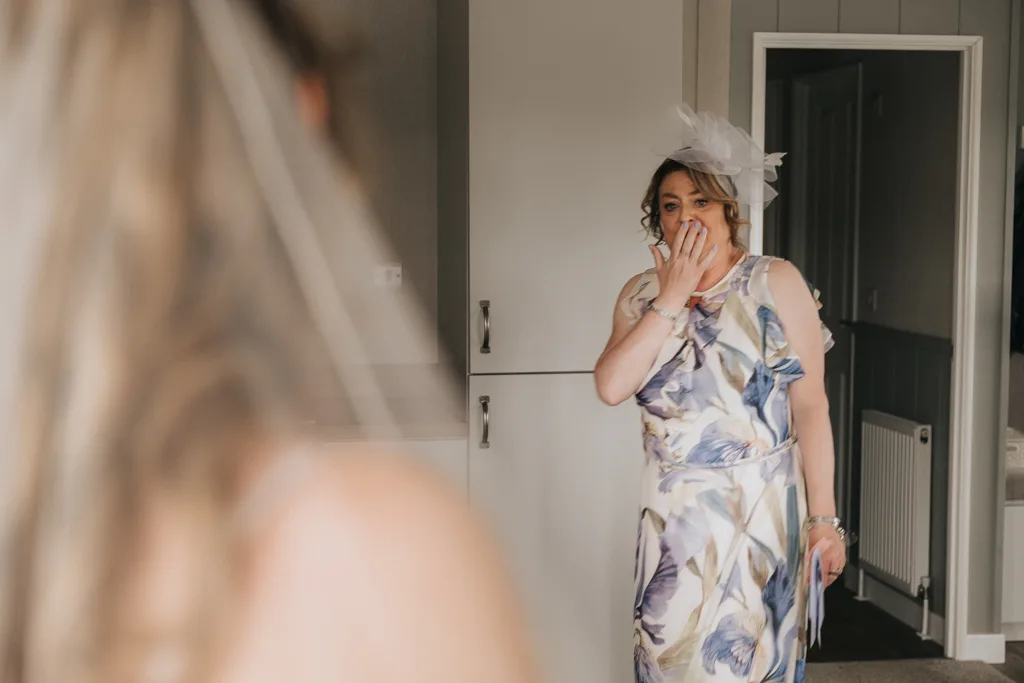 A woman in a floral dress and white fascinator covers her mouth in surprise, facing someone in a wedding dress. This emotional wedding photography moment unfolds indoors at Brackenborough, with soft light, gray walls, and white cabinets. © Aimee Lince Photography - Wedding photographer in Lincolnshire, Yorkshire & Nottinghamshire