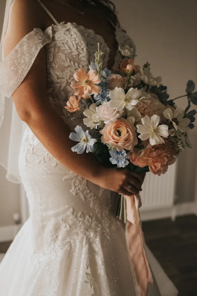 A bride in a beaded lace wedding gown holds a bouquet of pastel flowers, including pale pink roses and blue delphiniums, captured in soft indoor light—perfect for elegant wedding photography at Brackenborough. © Aimee Lince Photography - Wedding photographer in Lincolnshire, Yorkshire & Nottinghamshire
