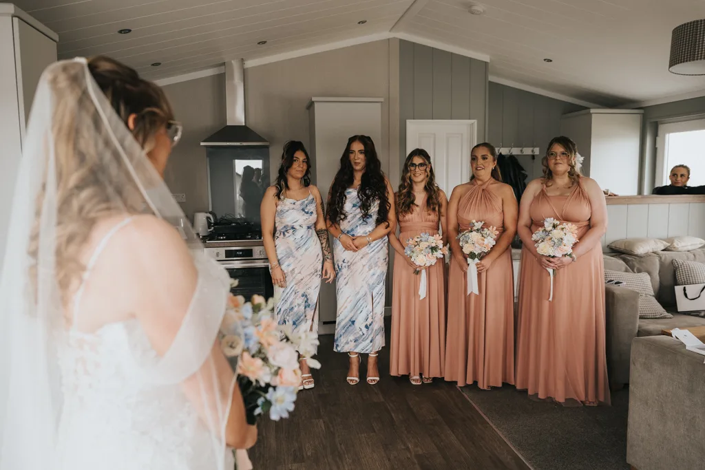 A bride in a white dress and veil stands with her back to the camera at a Brackenborough wedding, holding a bouquet. Four smiling bridesmaids—two in light blue patterned dresses and two in light brown—pose inside a modern kitchen-living room. © Aimee Lince Photography - Wedding photographer in Lincolnshire, Yorkshire & Nottinghamshire