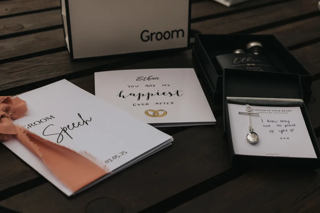 A flat lay of groom’s wedding items on a wooden table at Brackenborough: a “Groom Speech” booklet with peach ribbon, a heartfelt card, a gift box with necklace, and a box labeled “Groom” with two black flasks—perfect for photography. © Aimee Lince Photography - Wedding photographer in Lincolnshire, Yorkshire & Nottinghamshire