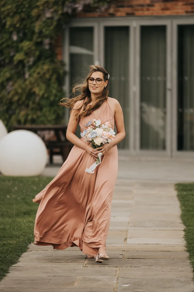 A woman in a flowing peach-colored dress walks along a stone path at Brackenborough, holding a bouquet of flowers. Her long hair blows in the wind as she passes glass doors and greenery—perfect for wedding photography. © Aimee Lince Photography - Wedding photographer in Lincolnshire, Yorkshire & Nottinghamshire