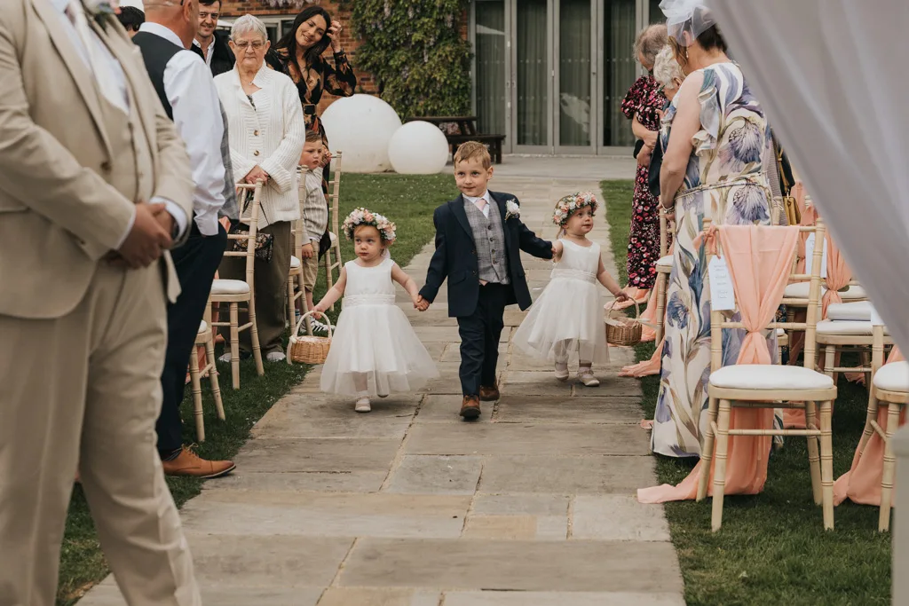 Three young children walk down the Brackenborough wedding aisle. Two flower girls in white dresses and flower crowns hold hands with a boy in a blue suit. Guests smile from their seats as photography captures this charming moment. © Aimee Lince Photography - Wedding photographer in Lincolnshire, Yorkshire & Nottinghamshire