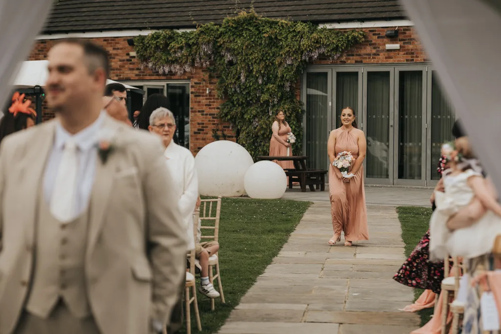 A bridesmaid in a blush pink dress walks down an outdoor wedding aisle holding a bouquet. Guests watch as brackenborough greenery climbs the brick building, with large white spheres on the lawn—a beautiful moment captured by photography. © Aimee Lince Photography - Wedding photographer in Lincolnshire, Yorkshire & Nottinghamshire
