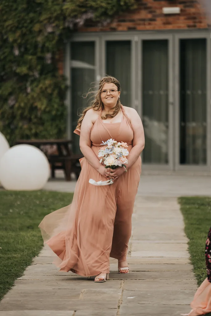 A woman in a flowing peach bridesmaid dress walks outside on a stone path at Brackenborough, holding a bouquet of flowers. Her hair is blown by the wind, with wedding photography capturing greenery and large windows in the background. © Aimee Lince Photography - Wedding photographer in Lincolnshire, Yorkshire & Nottinghamshire