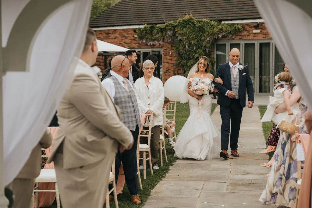 A smiling bride in a white dress holding a bouquet walks down an outdoor wedding aisle with an older man. Guests watch as white drapes frame the scene, and Brackenborough’s ivy-covered brick building stands in the background. © Aimee Lince Photography - Wedding photographer in Lincolnshire, Yorkshire & Nottinghamshire