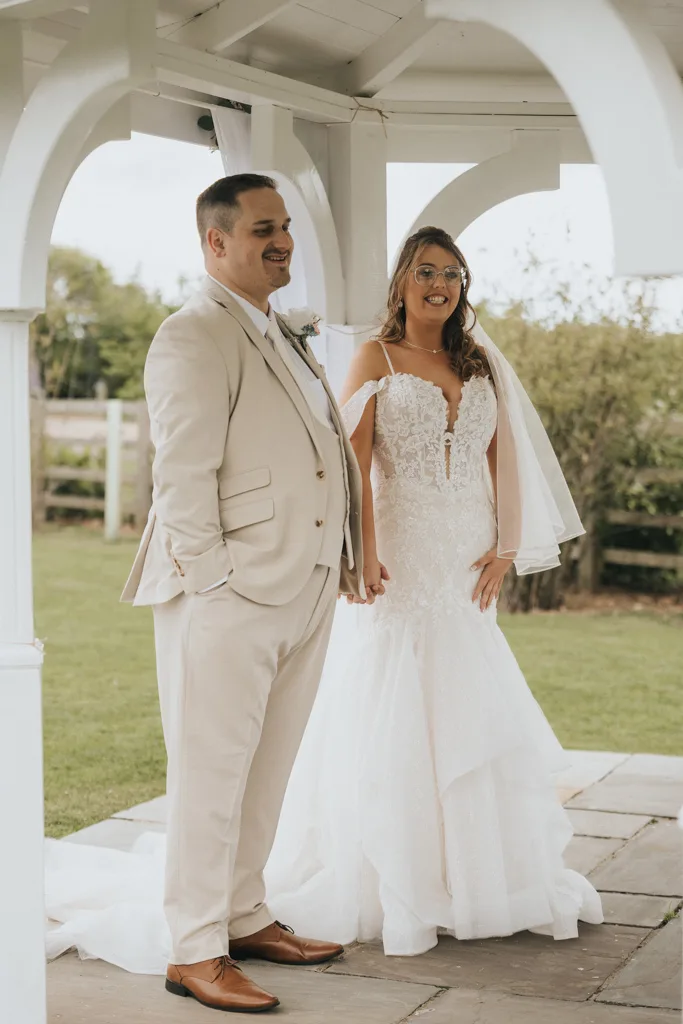 A bride and groom stand hand in hand under a white gazebo in this beautiful wedding photography at Brackenborough. The groom wears a beige suit; the bride dazzles in her off-the-shoulder lace gown, both smiling with green grass and a wooden fence behind them. © Aimee Lince Photography - Wedding photographer in Lincolnshire, Yorkshire & Nottinghamshire
