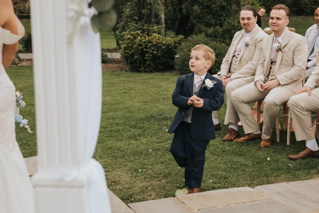 A young boy in a navy suit walks down an outdoor wedding aisle, looking up and smiling. Three men in light suits watch as the bride stands nearby. The lush garden scene is beautifully captured by Brackenborough wedding photography. © Aimee Lince Photography - Wedding photographer in Lincolnshire, Yorkshire & Nottinghamshire