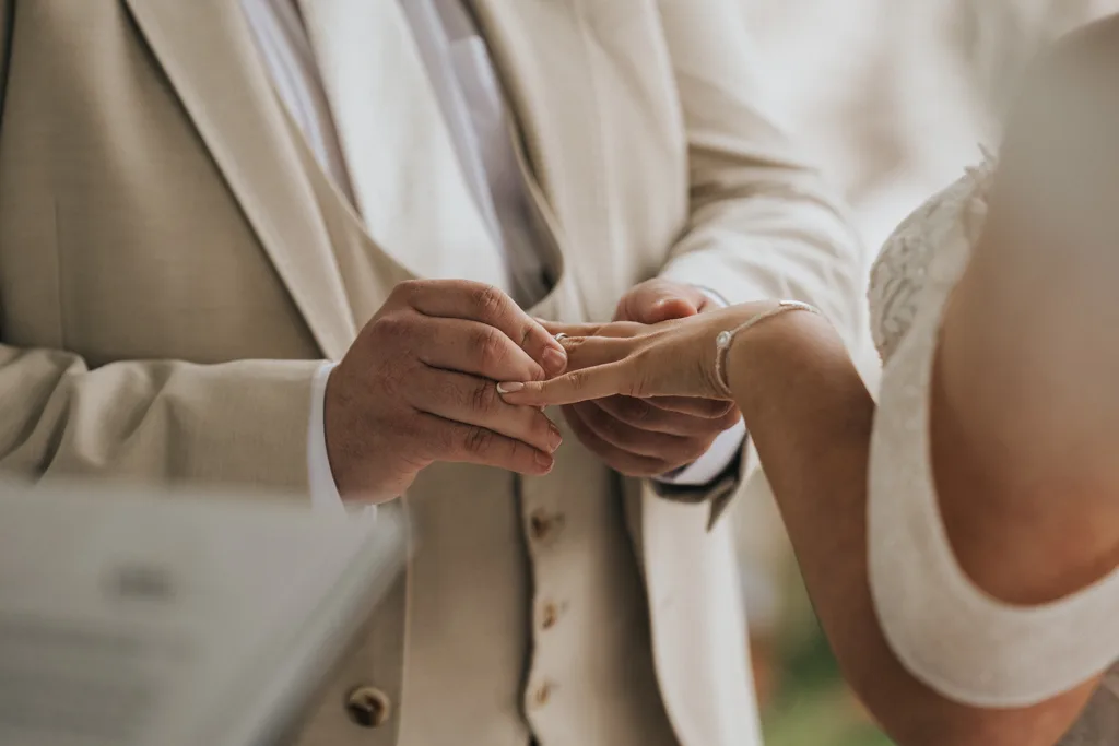 A close-up photo by Brackenborough wedding photography shows a groom in a beige suit gently placing a ring on the bride’s finger. The bride wears a white dress and delicate bracelet, with their hands in sharp focus against a softly blurred background. © Aimee Lince Photography - Wedding photographer in Lincolnshire, Yorkshire & Nottinghamshire
