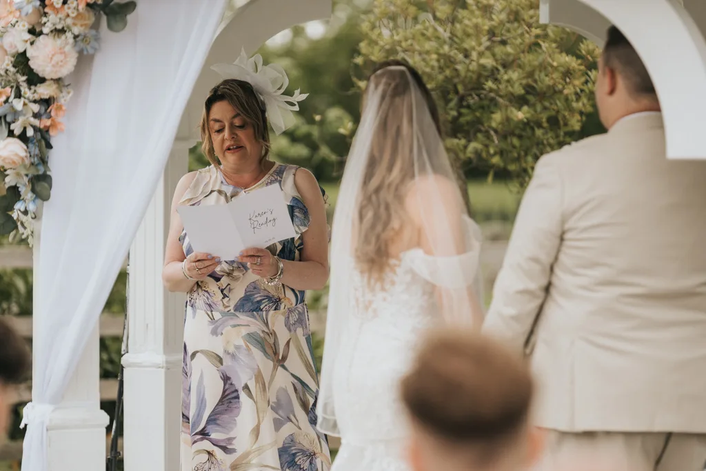 A woman in a floral dress reads from a sheet of paper under a white arch adorned with flowers at a wedding. She stands beside the bride and groom, with trees and greenery in the background—perfect for Brackenborough photography. © Aimee Lince Photography - Wedding photographer in Lincolnshire, Yorkshire & Nottinghamshire