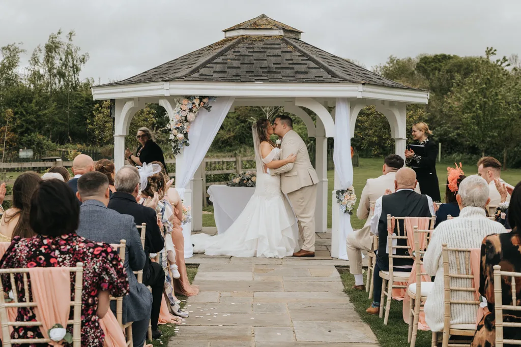 A bride and groom share a kiss under a decorated white gazebo at their outdoor wedding ceremony. Guests clap while Brackenborough’s lush greenery surrounds them—perfect for timeless photography moments. © Aimee Lince Photography - Wedding photographer in Lincolnshire, Yorkshire & Nottinghamshire