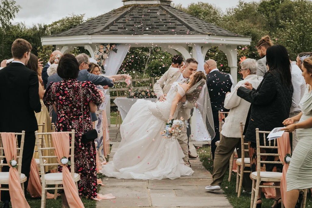 Brackenborough Lakes Resort Hotel Wedding Photos Louth Lincolnshire Chloe & Ethan 271 of 704 © Aimee Lince Photography - 03.05.2025 A newlywed couple shares a kiss as the groom dips the bride under a gazebo. Guests on decorated chairs toss flower petals in celebration, while Brackenborough wedding photography captures joyful expressions and lush greenery all around. © Aimee Lince Photography - Wedding photographer in Lincolnshire, Yorkshire & Nottinghamshire
