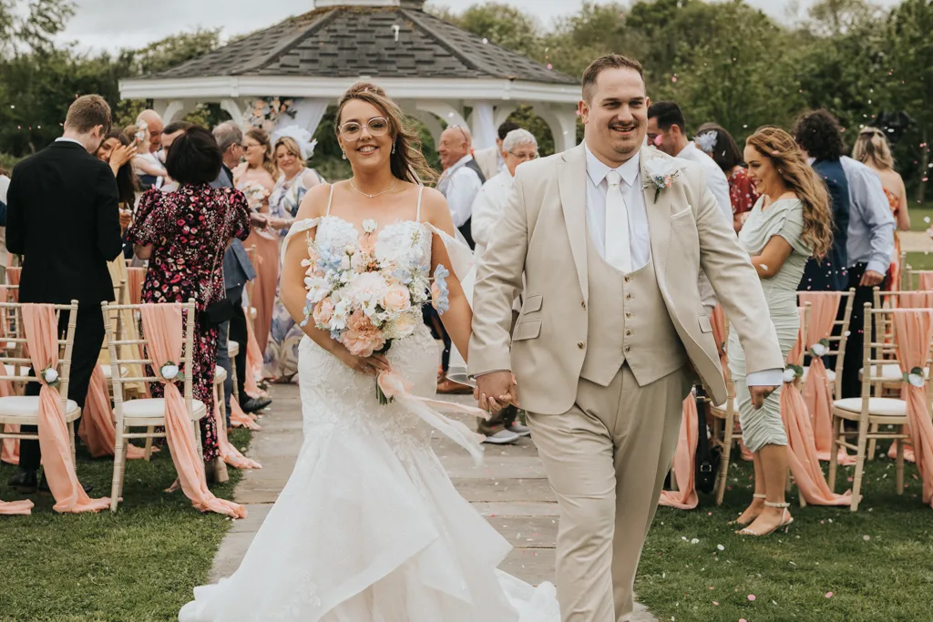 A bride and groom, smiling and holding hands, walk down the aisle outdoors after their Brackenborough wedding ceremony. The bride wears a white gown and holds a bouquet; the groom, in a beige suit, as guests celebrate beneath a gazebo—perfect for photography. © Aimee Lince Photography - Wedding photographer in Lincolnshire, Yorkshire & Nottinghamshire