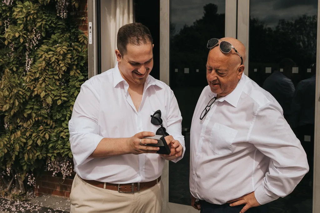 Two men in white shirts stand outdoors amid greenery, sharing a moment captured in brackenborough wedding photography. The younger man shows the older man a black box with sunglasses, both wearing shades and smiling warmly. © Aimee Lince Photography - Wedding photographer in Lincolnshire, Yorkshire & Nottinghamshire