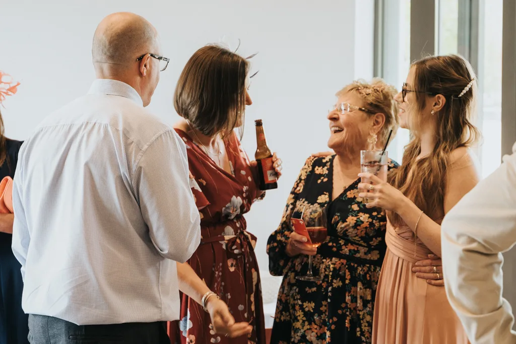 Four adults stand indoors at a Brackenborough wedding, talking and smiling. Two women wear floral dresses, one in red and one in black; another wears a peach dress. A bald man in glasses joins them as sunlight pours through large windows—a joyful moment of photography. © Aimee Lince Photography - Wedding photographer in Lincolnshire, Yorkshire & Nottinghamshire