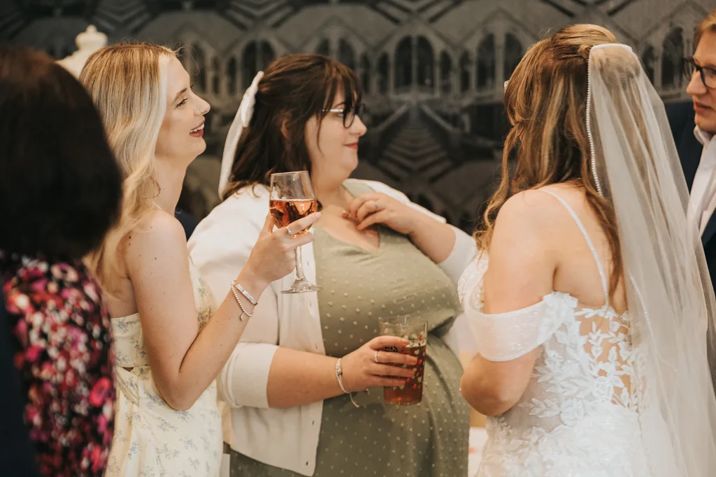 Three women at a wedding reception chat and smile. Captured by Brackenborough Photography, one bride in a white dress stands with her back turned while two guests in yellow and green dresses hold drinks, engaged in conversation. © Aimee Lince Photography - Wedding photographer in Lincolnshire, Yorkshire & Nottinghamshire
