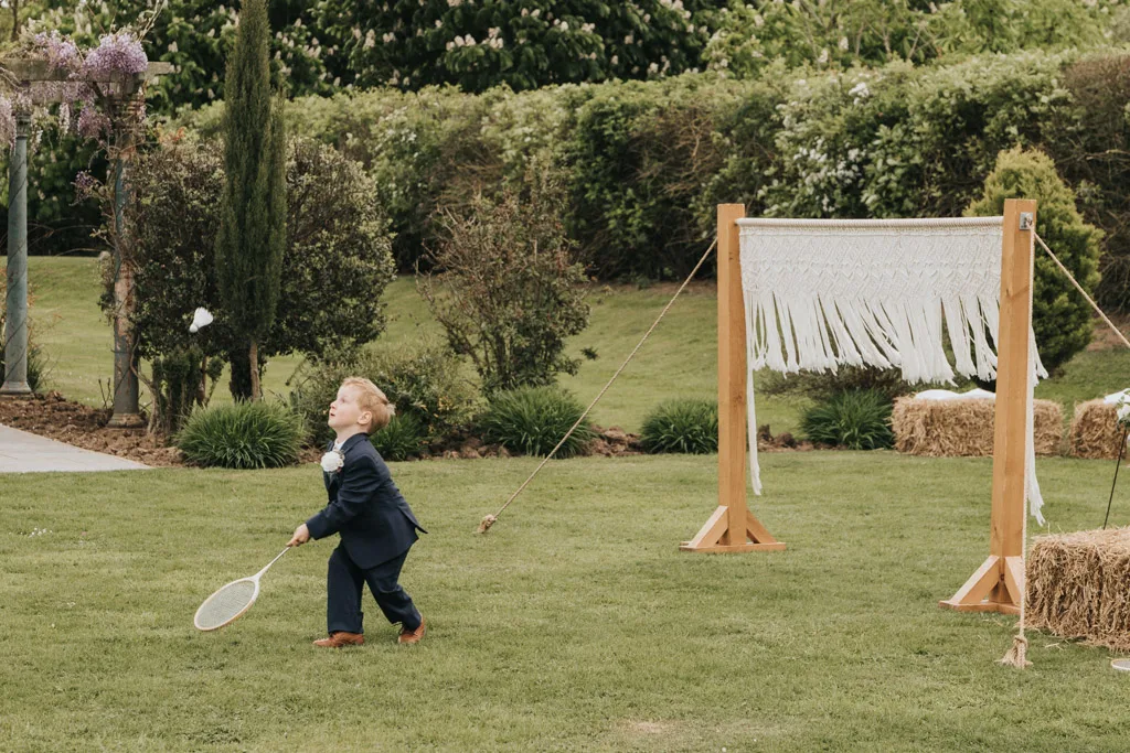 Brackenborough Lakes Resort Hotel Wedding Photos Louth Lincolnshire Chloe & Ethan 318 of 704 © Aimee Lince Photography - 03.05.2025 A young boy in a navy suit plays badminton outdoors at a Brackenborough wedding, swinging a racket at a shuttlecock. He stands on green grass near a rustic net, surrounded by bushes and hay bales—perfect for candid photography. © Aimee Lince Photography - Wedding photographer in Lincolnshire, Yorkshire & Nottinghamshire