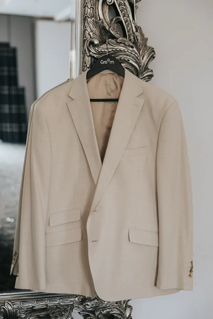 A beige suit jacket hangs on a black “Groom” hanger, displayed on an ornate silver-framed mirror. The softly lit background and elegant composition capture the anticipation of a wedding through refined photography. © Aimee Lince Photography - Wedding photographer in Lincolnshire, Yorkshire & Nottinghamshire