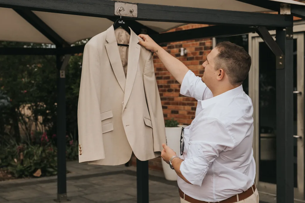 A man in a white shirt holds a beige suit jacket on a black hanger outdoors at Brackenborough. He examines the jacket under a pergola, with greenery, glass doors, and brick wall behind—a perfect moment of wedding photography. © Aimee Lince Photography - Wedding photographer in Lincolnshire, Yorkshire & Nottinghamshire