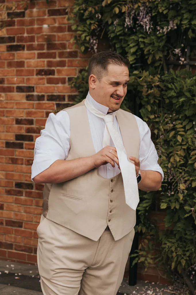 A man in a beige vest and pants stands outdoors at Brackenborough, adjusting a white necktie. With short dark hair and a slight smile, he poses for wedding photography by a brick wall and leafy greenery, wearing a white shirt with rolled-up sleeves. © Aimee Lince Photography - Wedding photographer in Lincolnshire, Yorkshire & Nottinghamshire