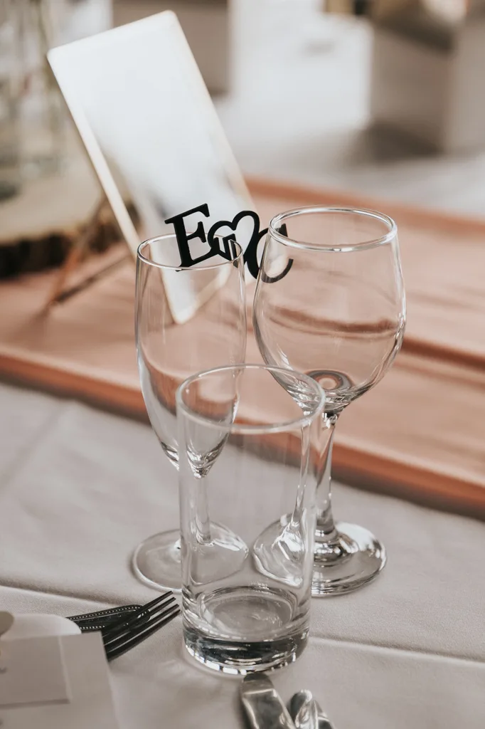 Three empty glasses—a wine glass, a flute, and a tumbler—are arranged on a white tablecloth at a Brackenborough wedding. Behind them, a decorative monogram with “E” and a heart is visible, captured through elegant photography. © Aimee Lince Photography - Wedding photographer in Lincolnshire, Yorkshire & Nottinghamshire
