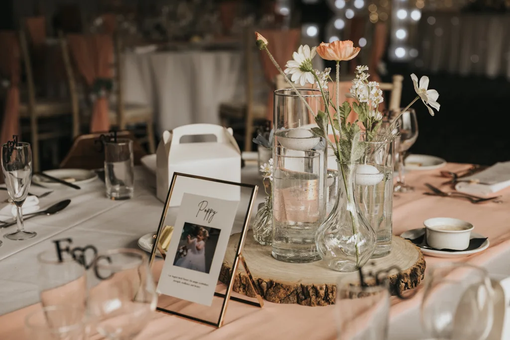 A wedding table centerpiece at Brackenborough features glass vases with flowers and floating candles on a wooden slice. A framed photo, perfect for photography, sits beside a name card, elegant place settings, champagne glasses, and a white favor box. © Aimee Lince Photography - Wedding photographer in Lincolnshire, Yorkshire & Nottinghamshire