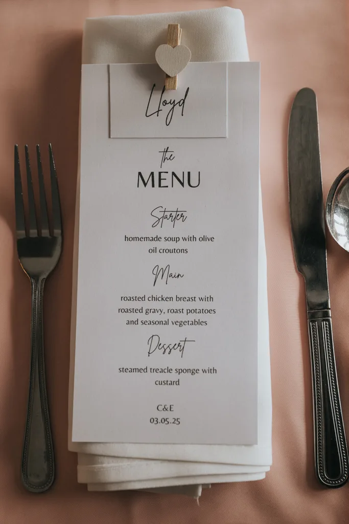 A wedding menu card labeled "Lloyd" is clipped with a small heart peg to a folded white napkin. Captured by Brackenborough photography, the menu sits between a fork and knife on a soft pink tablecloth, listing soup, chicken, and treacle sponge. © Aimee Lince Photography - Wedding photographer in Lincolnshire, Yorkshire & Nottinghamshire