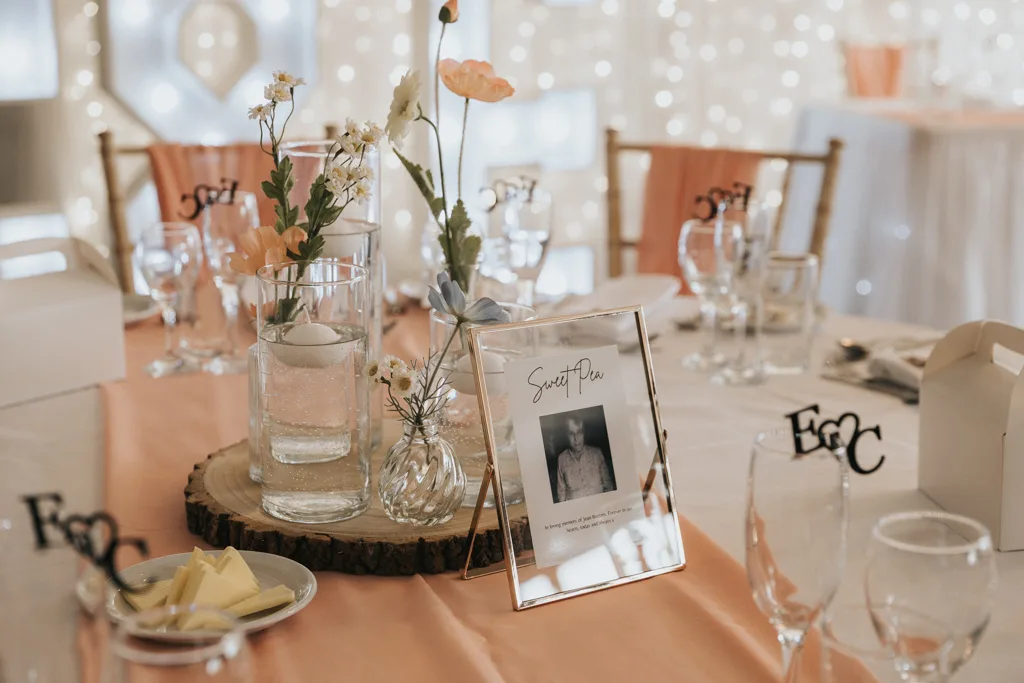 Brackenborough Lakes Resort Hotel Wedding Photos Louth Lincolnshire Chloe & Ethan 446 of 704 © Aimee Lince Photography - 03.05.2025 A round table set for a wedding at Brackenborough features a peach table runner, floral centerpieces in glass vases, a framed “Sweet Pea” photo—perfect for photography. Place settings, fairy lights, and peach-draped chairs complete the elegant scene. © Aimee Lince Photography - Wedding photographer in Lincolnshire, Yorkshire & Nottinghamshire