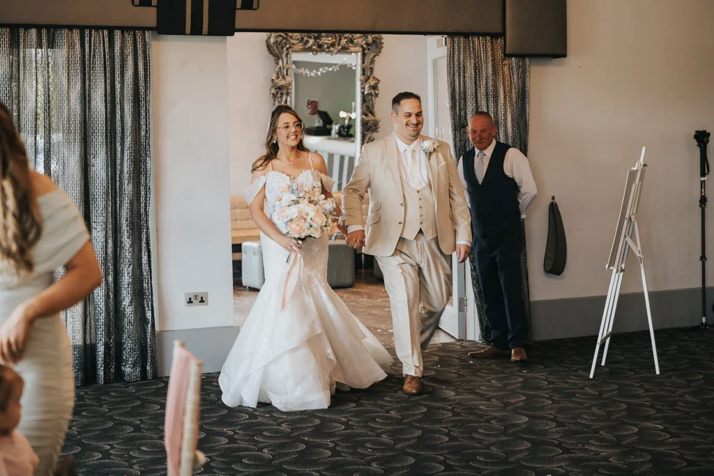 Brackenborough Lakes Resort Hotel Wedding Photos Louth Lincolnshire Chloe & Ethan 456 of 704 © Aimee Lince Photography - 03.05.2025 A bride in a white, off-shoulder gown and a groom in a tan suit walk arm-in-arm into their wedding at Brackenborough, smiling. The bride holds a bouquet of flowers. Guests and a man in a vest look on as photography captures the joyful moment. © Aimee Lince Photography - Wedding photographer in Lincolnshire, Yorkshire & Nottinghamshire