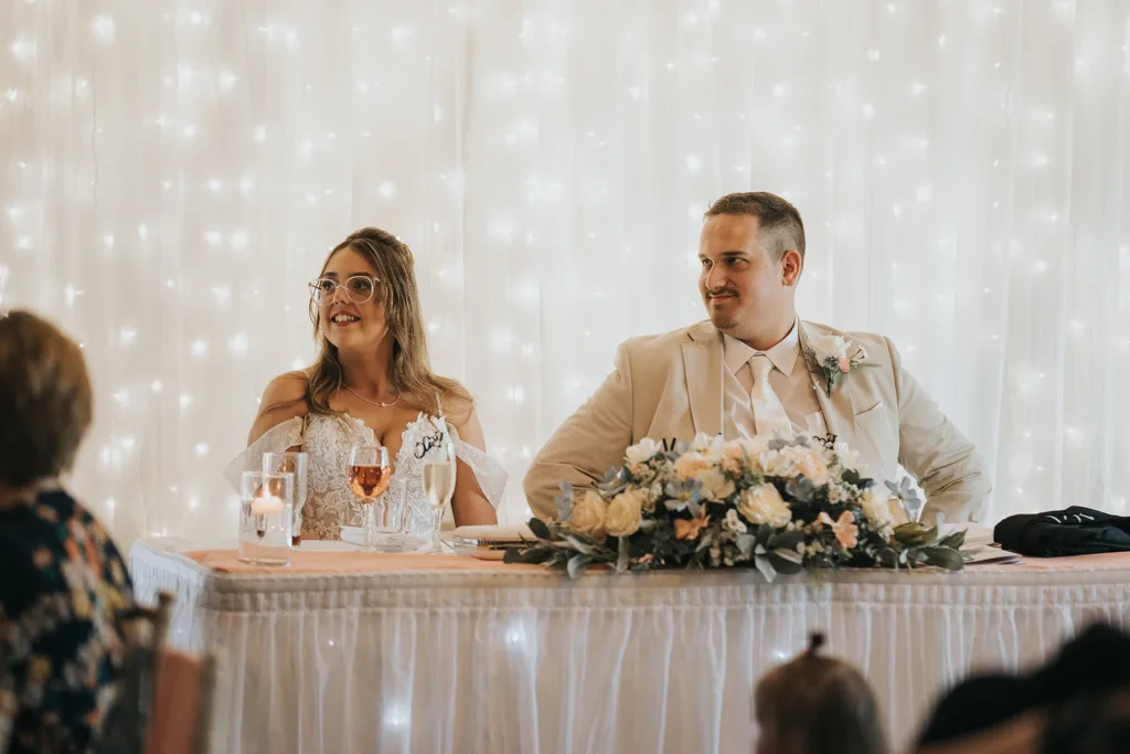A bride in a white dress and glasses and a groom in a beige suit sit at a beautifully decorated wedding table with flowers and candles, smiling. Behind them, twinkling fairy lights add magic to this Brackenborough photography moment. © Aimee Lince Photography - Wedding photographer in Lincolnshire, Yorkshire & Nottinghamshire
