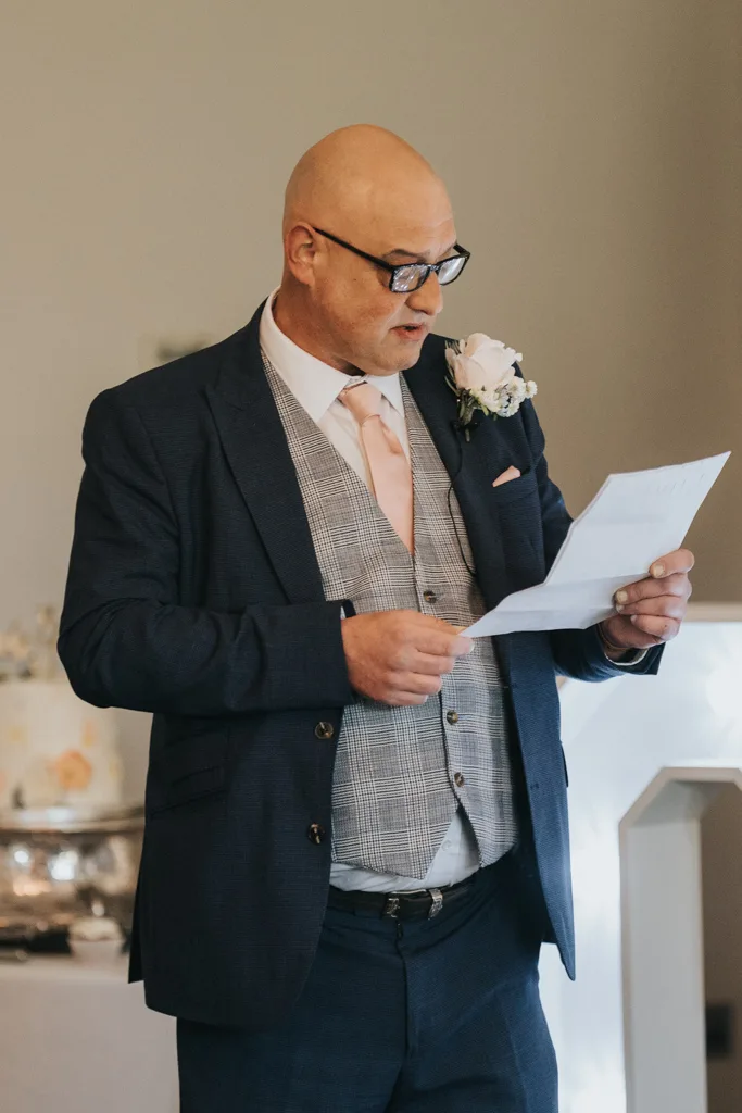 A bald man in glasses, wearing a navy blue suit, checked vest, and pink tie, stands indoors reading from a sheet of paper at a Brackenborough wedding. A decorated cake and silver dishes are softly focused in the background of this photography. © Aimee Lince Photography - Wedding photographer in Lincolnshire, Yorkshire & Nottinghamshire